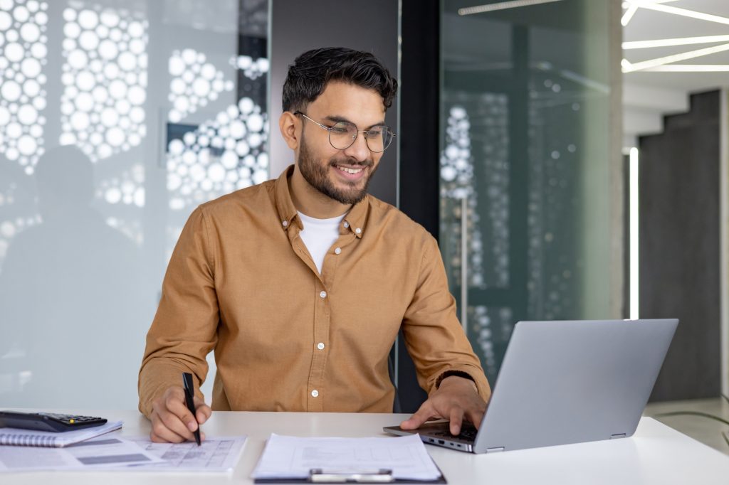 Smiling Indian man working on a laptop in the office, sitting at a table in front of a screen